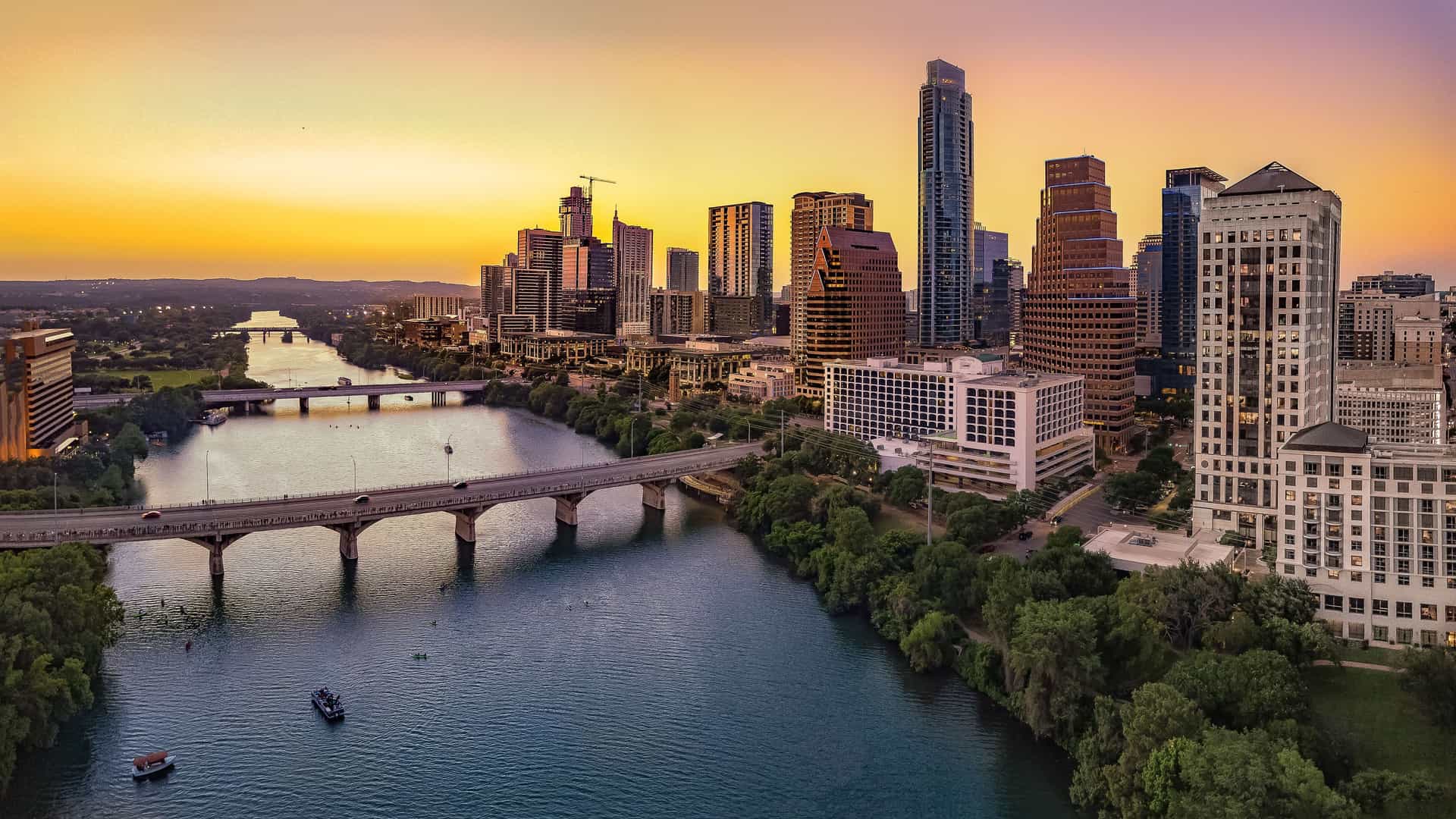 A scenic panoramic view of the Austin, Texas skyline at sunset. The Colorado River (also known as Lady Bird Lake) flows through the center, with the Congress Avenue Bridge crossing in the foreground. Modern skyscrapers line the riverbank, and the sky is painted with vibrant shades of orange, yellow, and purple.