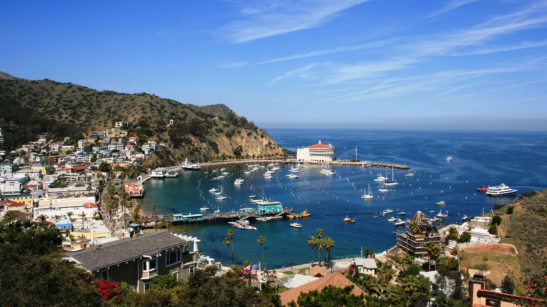 A high-angle view of Avalon, a beautiful harbor town on Santa Catalina Island, California, with boats dotting the bay and the iconic Catalina Casino building on a sunny day.