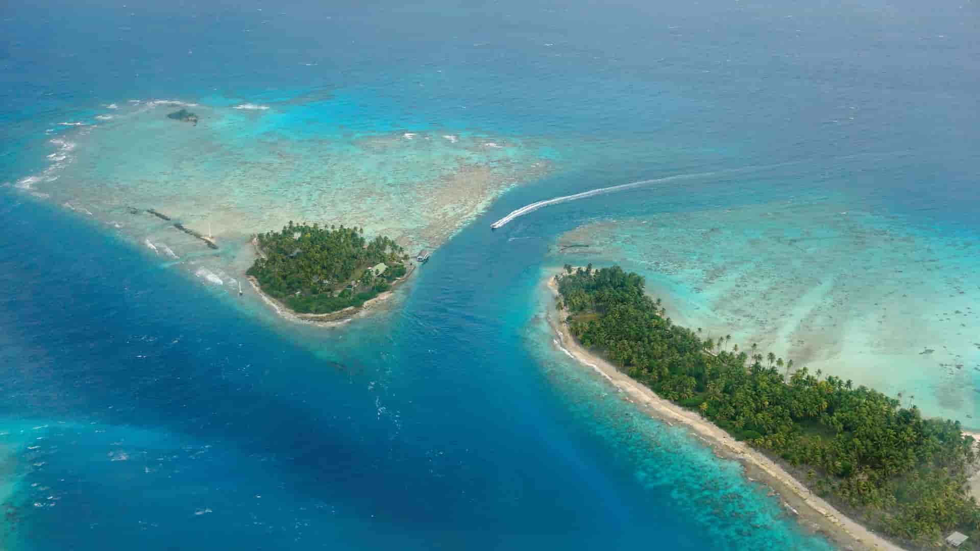 An aerial view of Avatoru, a small tropical island in Rangiroa, French Polynesia, showing a small boat navigating the channel between two landmasses surrounded by coral reefs.