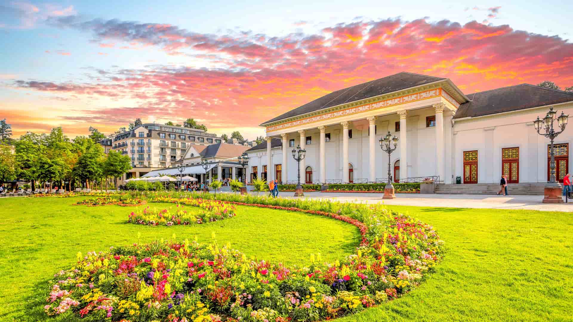 A picturesque view of the Kurhaus in Baden-Baden, Germany, a grand, white neoclassical building with columns. A vibrant green lawn with a long, curving bed of colorful flowers fills the foreground, all under a dramatic sunset sky with hues of orange, pink, and purple.