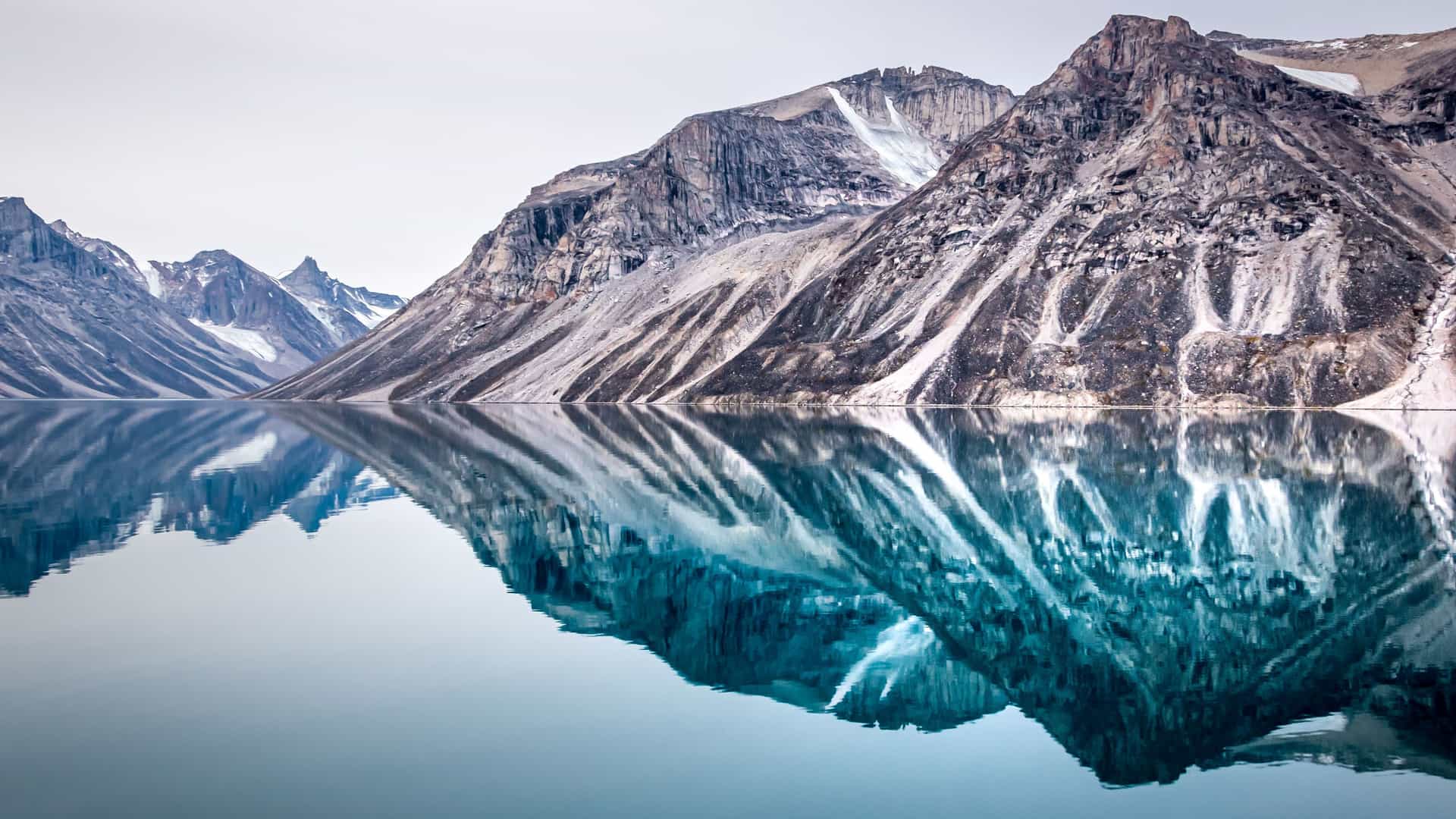 A breathtaking view of dramatic, rugged mountains in Baffin Island, Canada, with snow and striated rock on their slopes. The mountains are perfectly reflected in the still, glassy surface of a large body of water, creating a serene and symmetrical natural landscape under a pale sky.