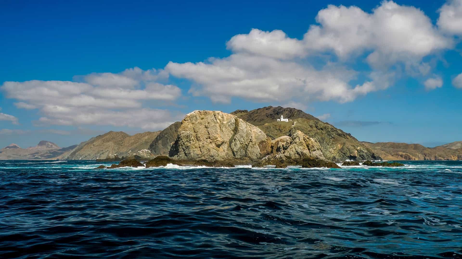 A panoramic view of an uninhabited island in Bahía Magdalena, Mexico, showing a rugged, rocky coastline, dark blue water, and a lighthouse perched on a high peak under a cloudy blue sky.