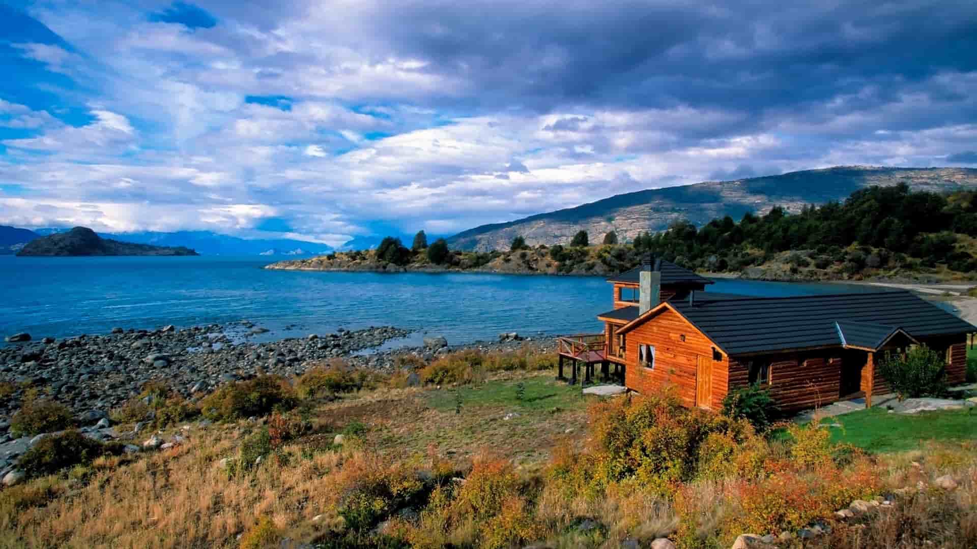 A rustic wooden lodge with a dark roof sits on the rocky shore of Baker Canal, a scenic waterway in the Patagonian region of Chile, with a mountainous landscape in the distance.
