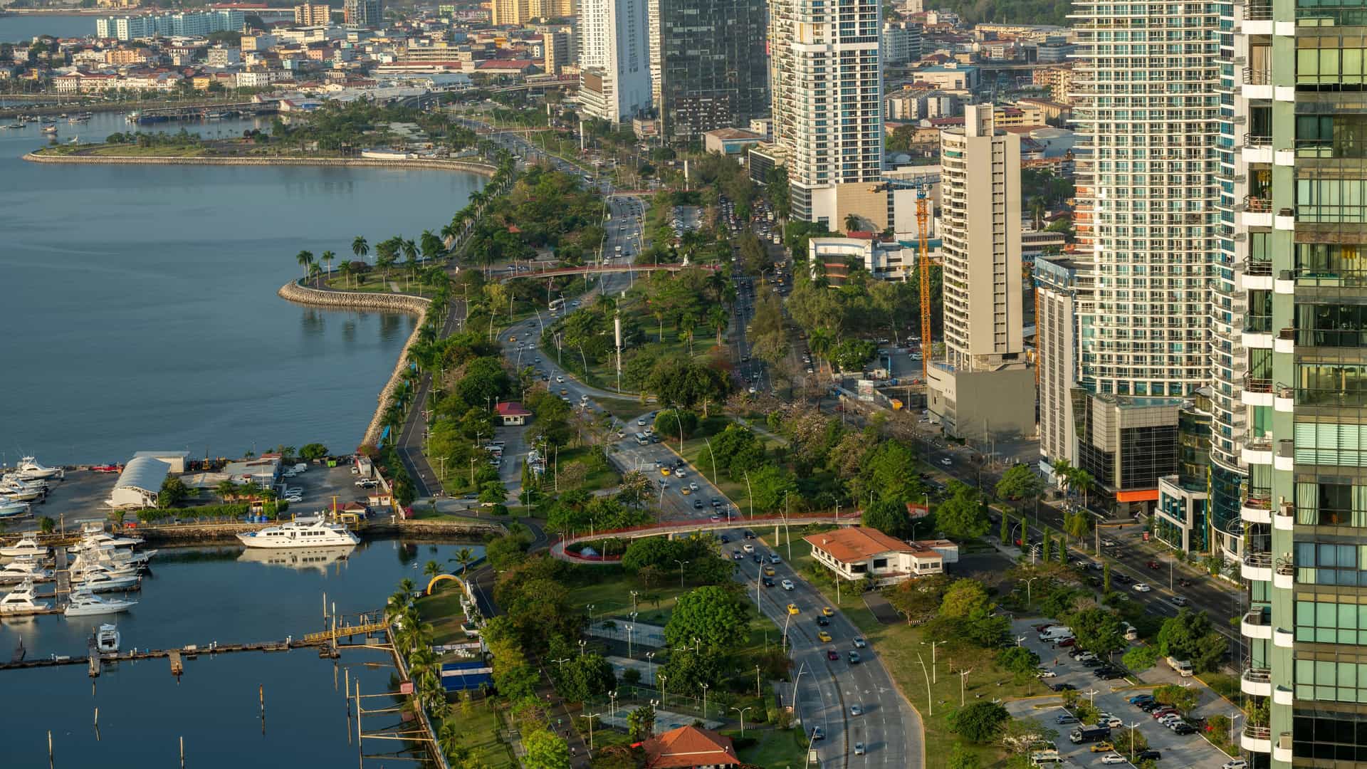 An aerial view of the bustling city of Balboa, Panama, with a scenic coastal road alongside a marina and tall modern skyscrapers in the background.