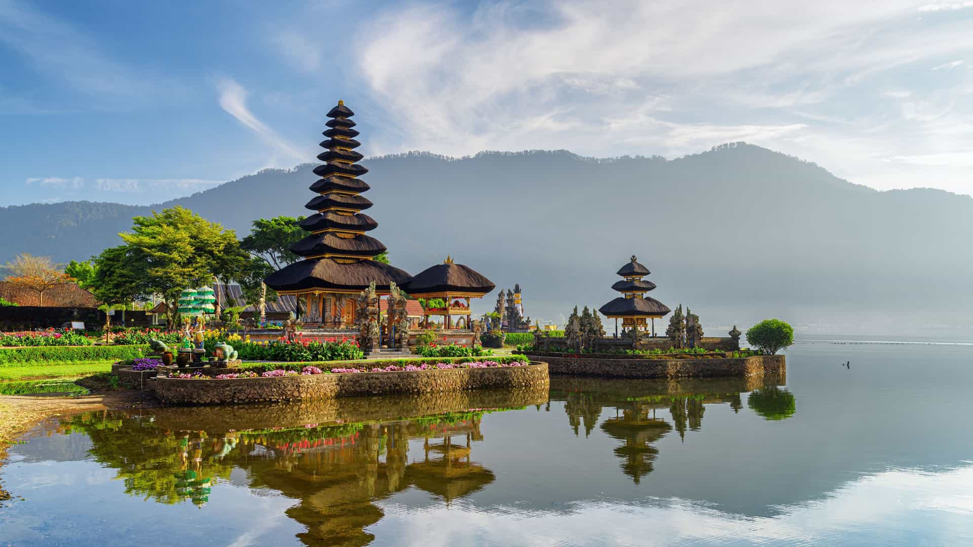A scenic view of the Ulun Danu Beratan Temple, a Hindu temple on the shores of Lake Beratan in Bali, with tiered pagodas reflecting in the calm water and mountains in the background.