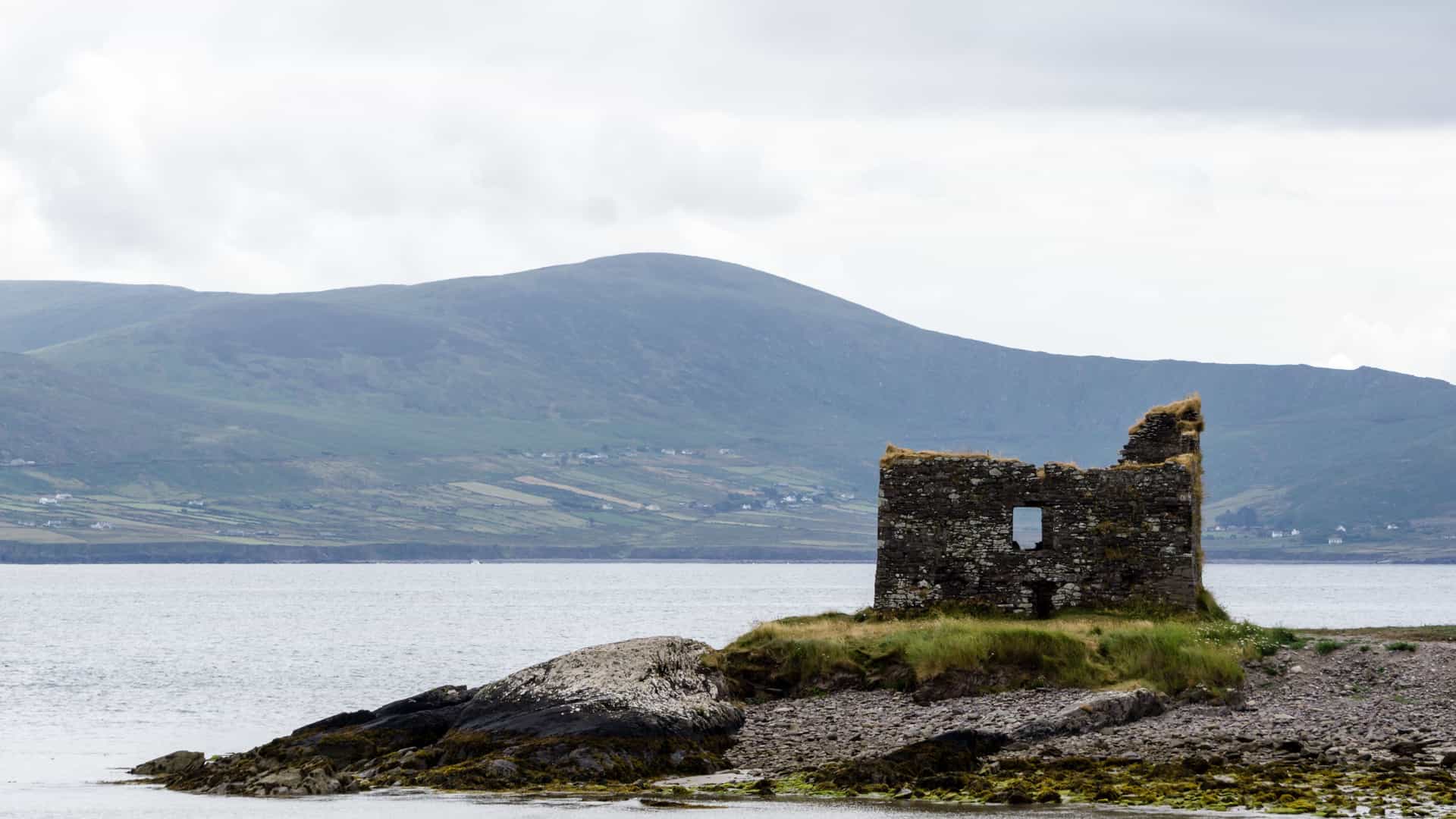A coastal landscape in Ballinskelligs featuring the ruins of an old stone structure on a rocky shoreline, surrounded by grassy patches and calm water, with rolling green hills and scattered buildings under a cloudy sky.