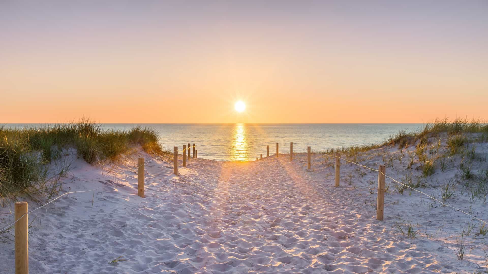 A sandy pathway with wooden posts and a rope fence leads over grassy dunes to the Baltic Sea as the sun sets over the horizon.