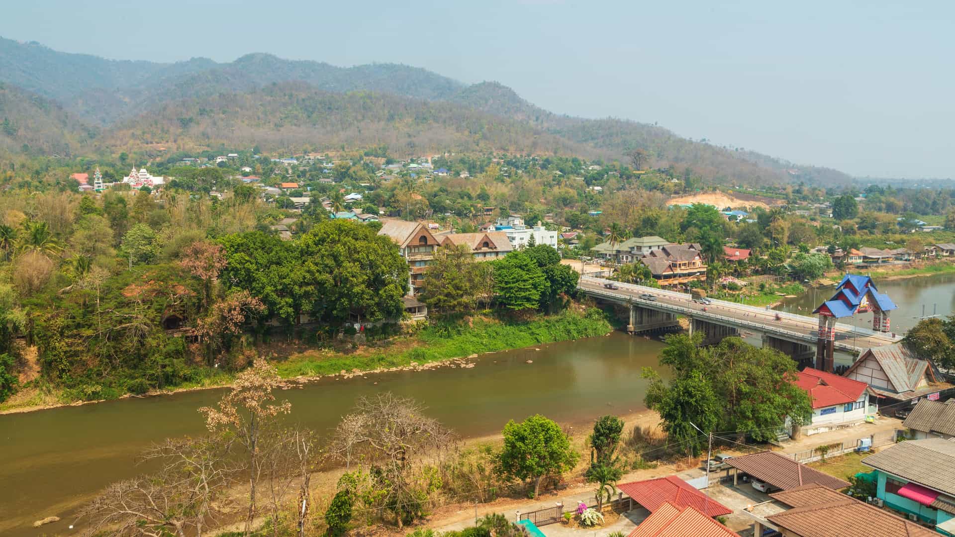 An elevated view of the town of Ban Tha Ton, Thailand, nestled between a wide, brown river and rolling, tree-covered hills. A concrete bridge crosses the river, connecting the two sides of the town, which is composed of a mix of houses with red and brown roofs.