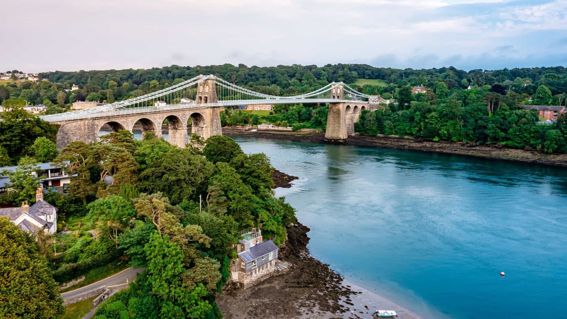 An aerial view of the historic Menai Suspension Bridge in Bangor, Wales, spanning a scenic waterway surrounded by lush green forests and homes on the shoreline.