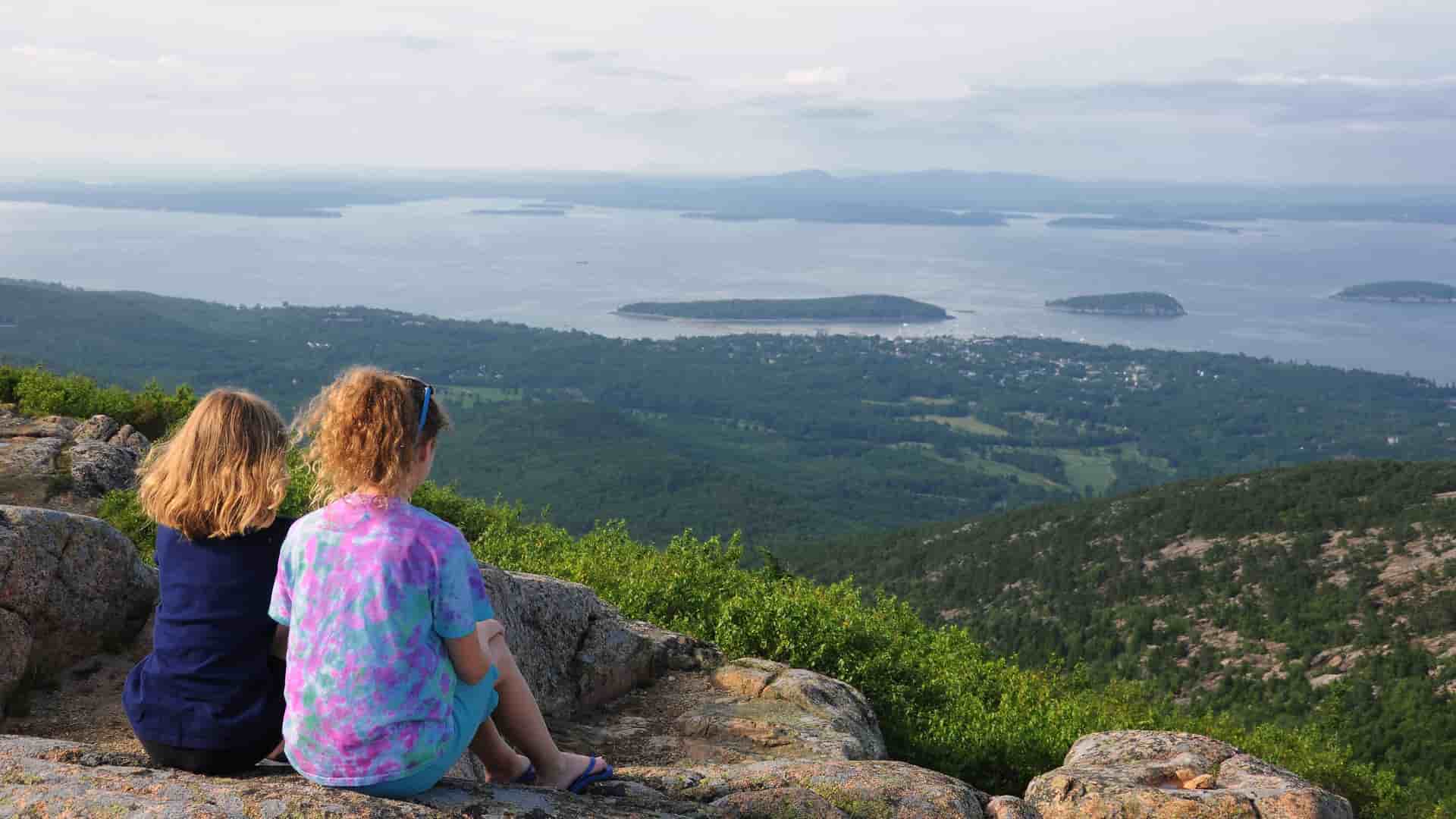 Two people sit on a rocky outcrop, enjoying a scenic view of Bar Harbor, Maine, with islands scattered across a calm bay and a vast forest below.