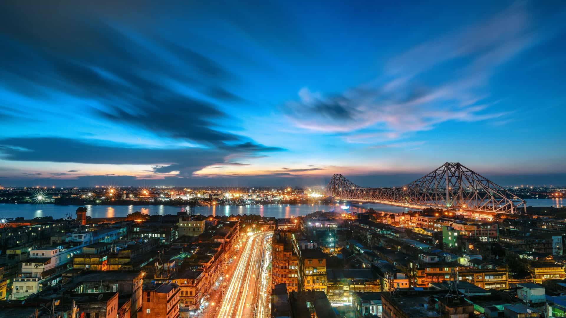 A breathtaking long-exposure city view of Baranagar in West Bengal, India, at dusk, with the illuminated Howrah Bridge and the Hooghly River in the background.