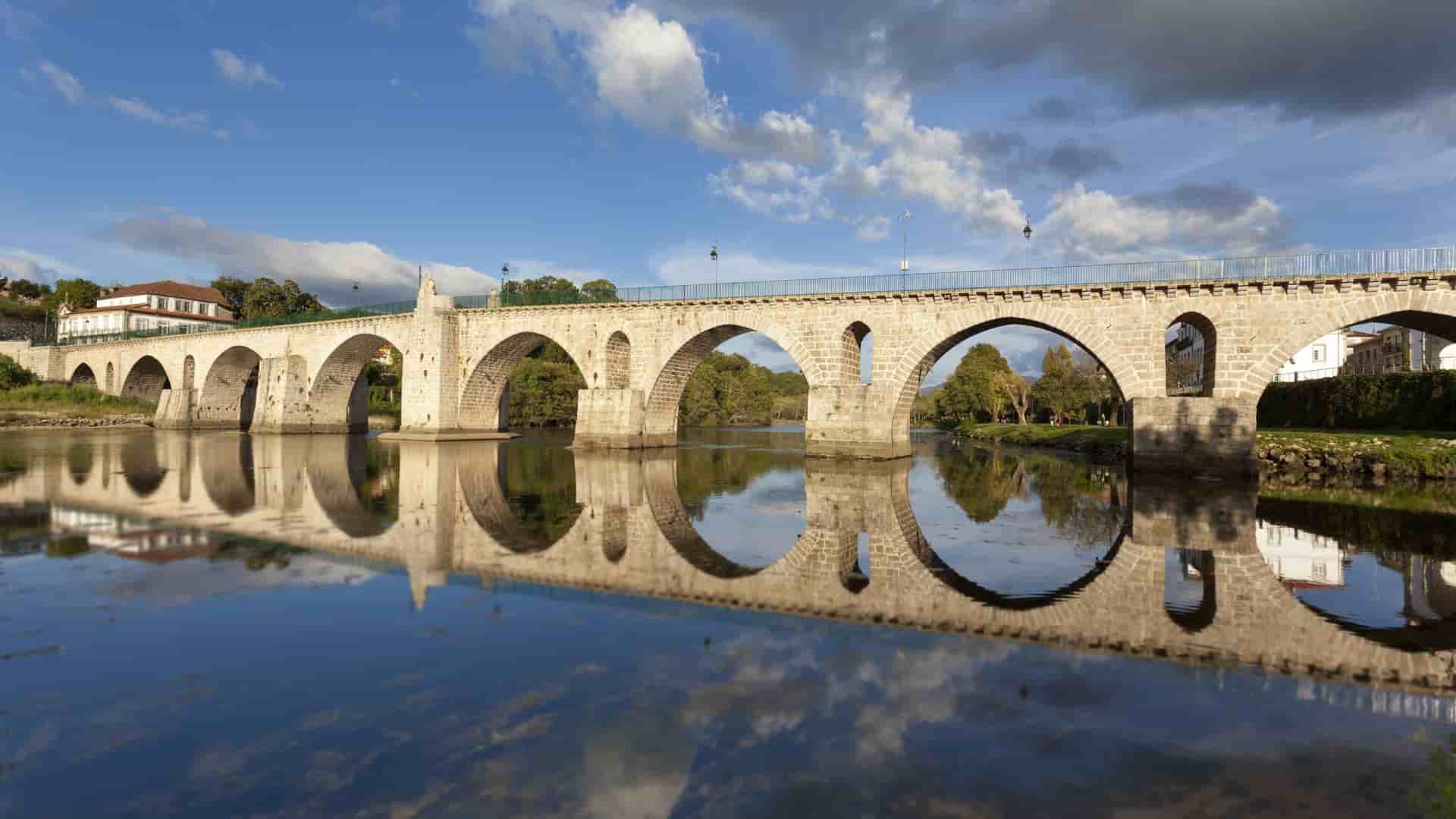 A scenic view of a Roman arch bridge spanning a calm river in Barca d'Alva, Portugal, with the bridge's arches and the surrounding landscape perfectly reflected in the still water below.