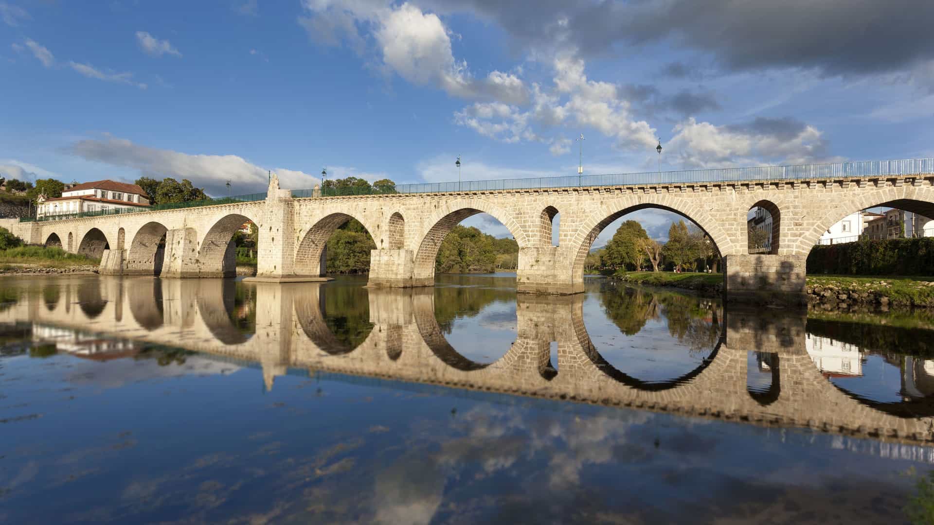 A scenic view of a Roman arch bridge spanning a calm river in Barca d'Alva, Portugal, with the bridge's arches and the surrounding landscape perfectly reflected in the still water below.