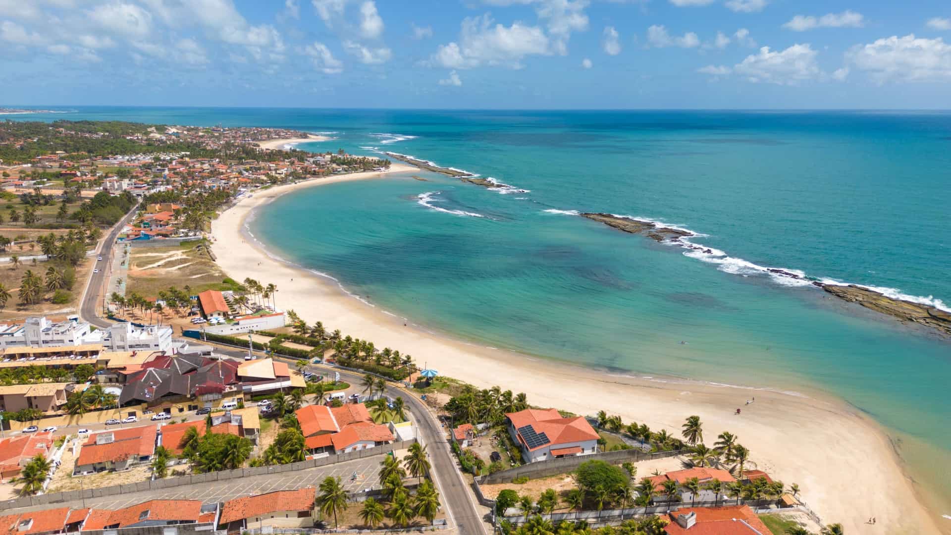 An aerial view of Barra do Jacuípe beach where the river meets the Atlantic Ocean, featuring a long coastline with a sandy beach and a small coastal town.