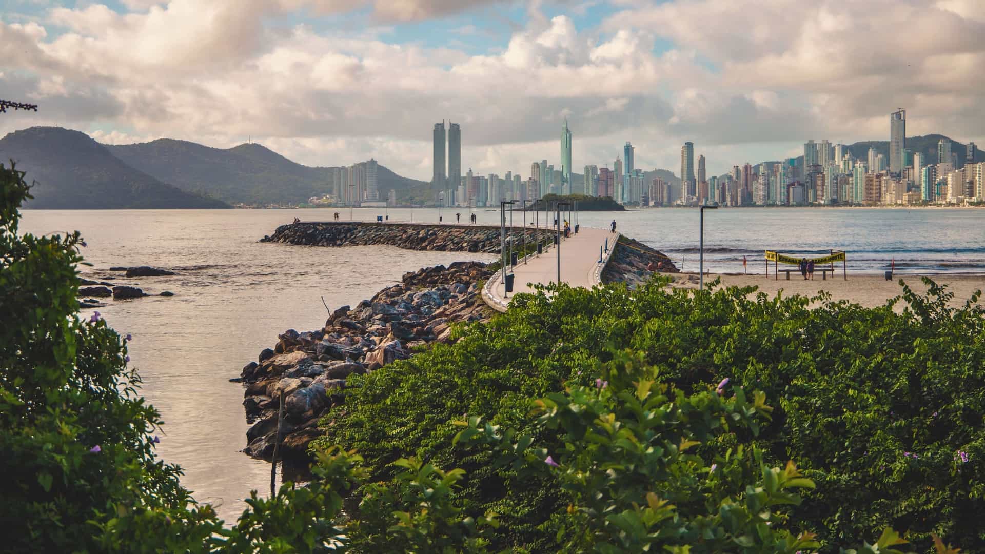 A long pedestrian walkway on a breakwater extending into the ocean, with the Balneário Camboriú skyline and a mountain in the background.