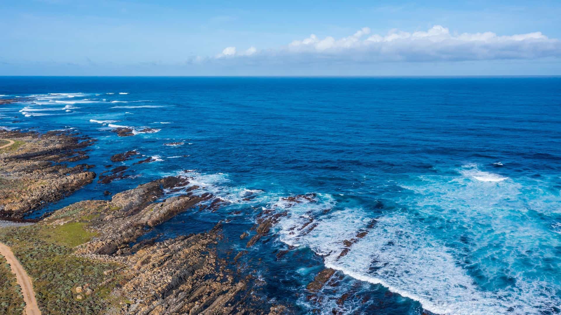An aerial view of the rugged coastline of the Bass Strait in Australia, featuring waves crashing against jagged rocks and a narrow dirt path along the shore.