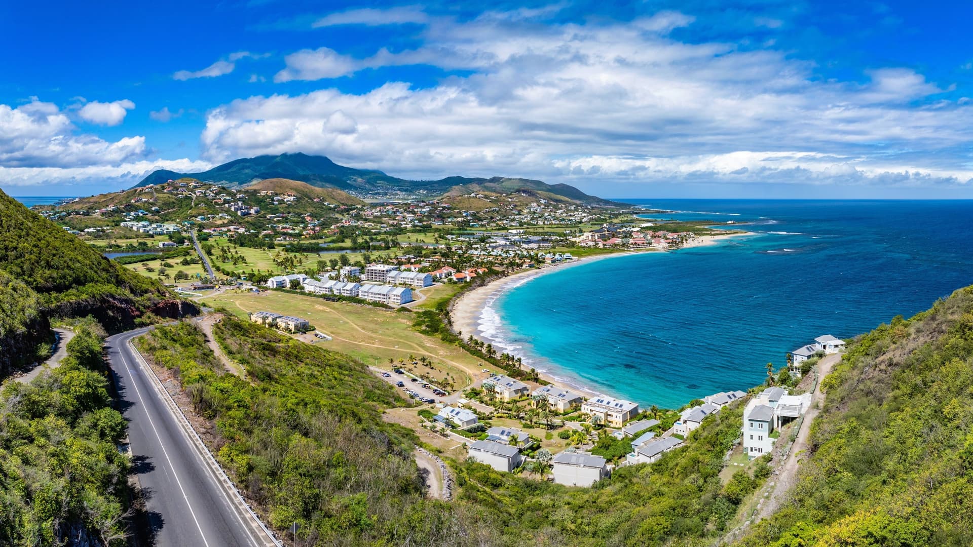 An aerial panoramic view of a winding road overlooking Frigate Bay and the Caribbean Sea, with rolling green hills, a beach, and the town of Basseterre, St. Kitts.