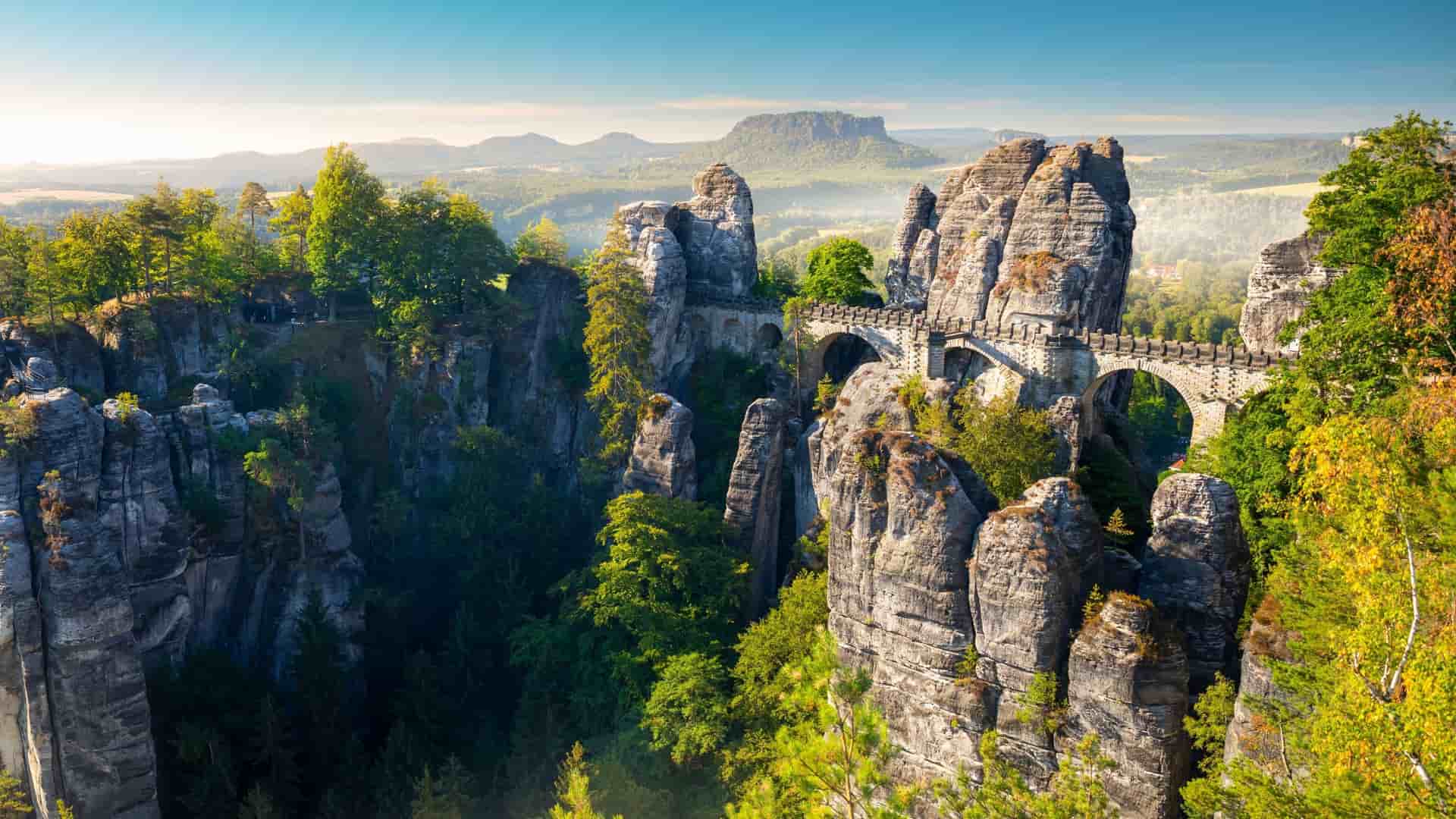 A dramatic landscape of the Bastei Bridge, a historic stone bridge with multiple arches connecting towering rock formations and jagged cliffs in Saxon Switzerland National Park, Germany.