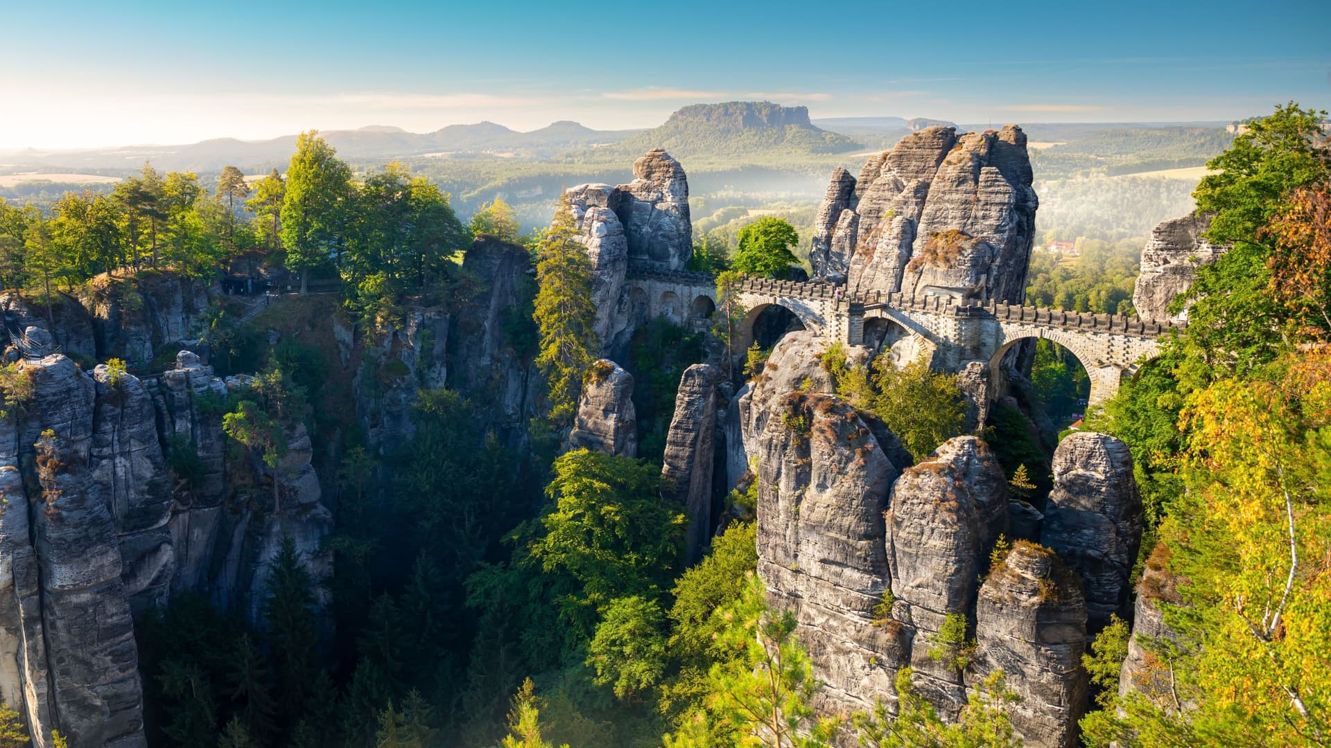 A dramatic landscape of the Bastei Bridge, a historic stone bridge with multiple arches connecting towering rock formations and jagged cliffs in Saxon Switzerland National Park, Germany.