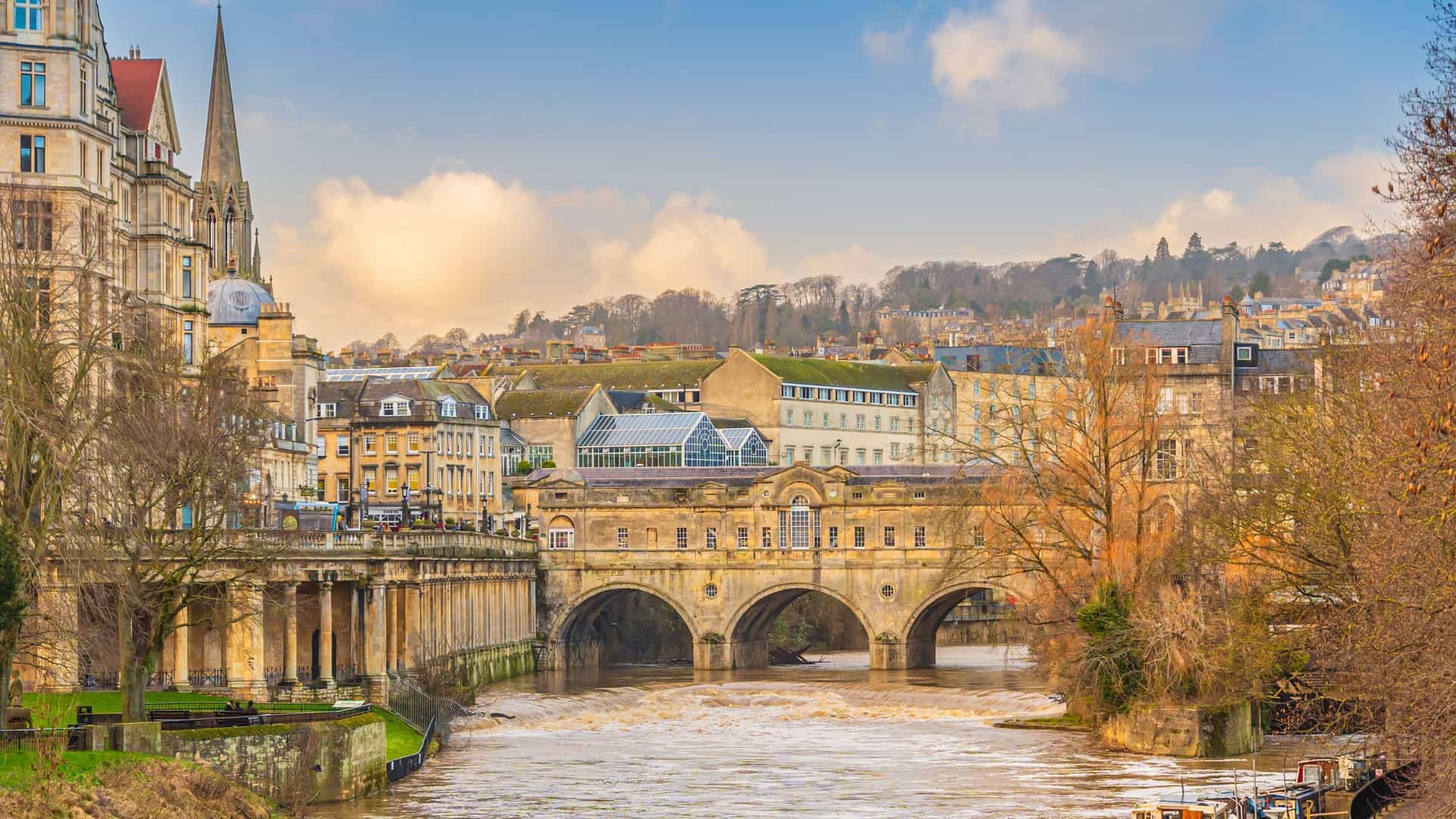 A view of the historic Pulteney Bridge and the River Avon in Bath, England, featuring the city's famous Georgian architecture and a church steeple in the background.