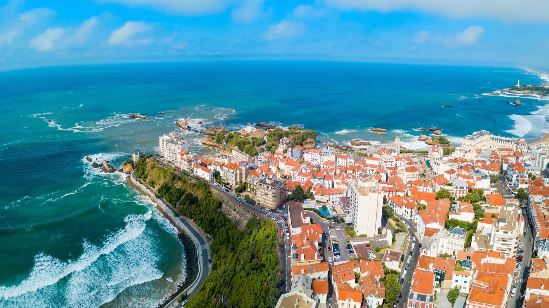 An aerial panoramic view of the Bay of Biscay coastline, showing the red-roofed buildings of Biarritz, France, along the ocean with waves crashing onto a rocky beach.