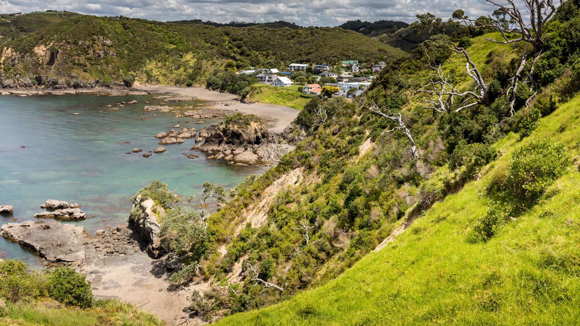 A panoramic view of the Bay of Islands, New Zealand, showing a small coastal town and a tranquil bay with rocky shorelines and lush green hills under a cloudy sky.