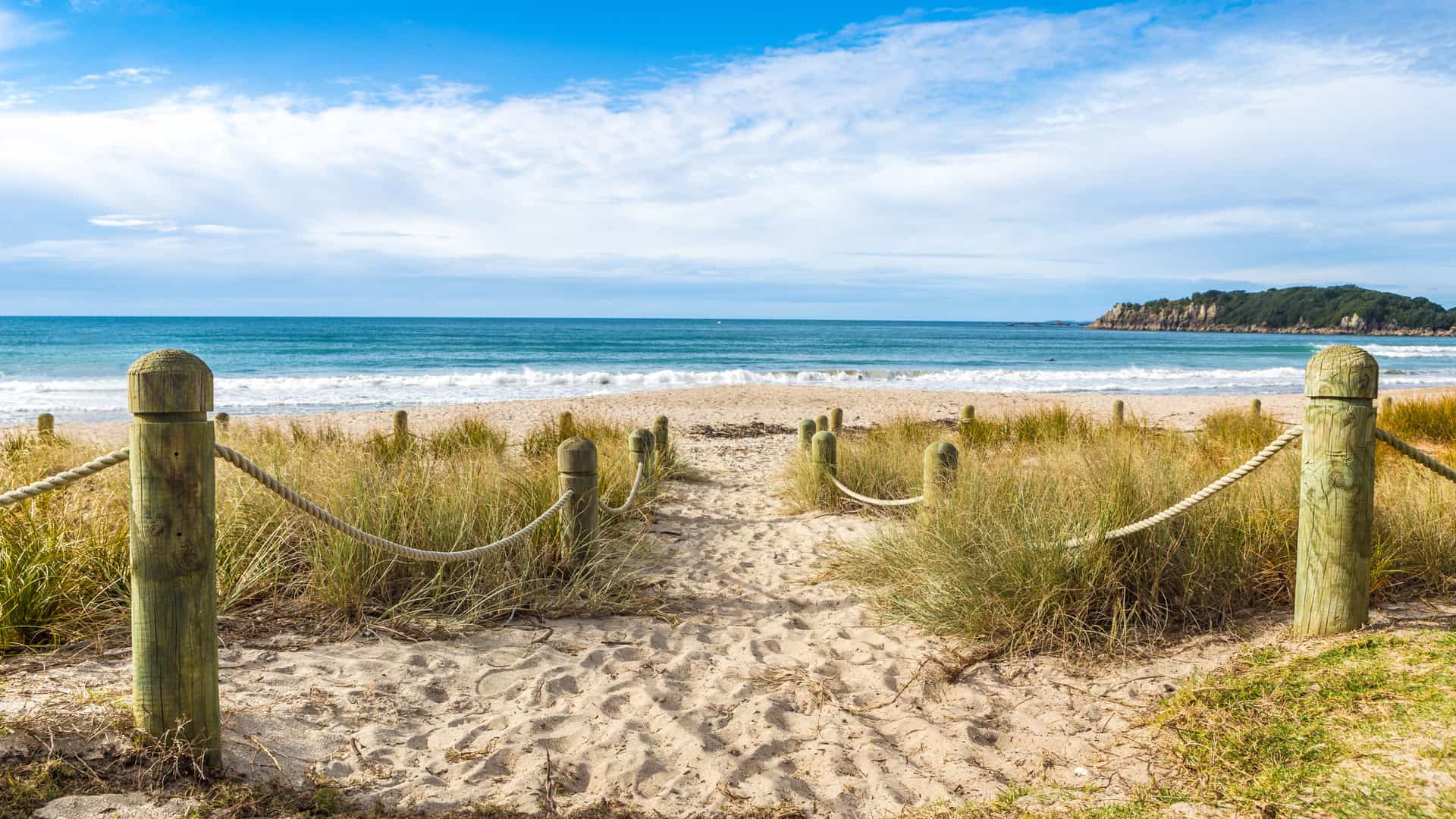 A sandy coastal pathway in Bay of Plenty bordered by rope‑lined wooden posts, leading through tall beach grass toward a calm blue sea, with a forested headland in the distance beneath a partly cloudy sky.