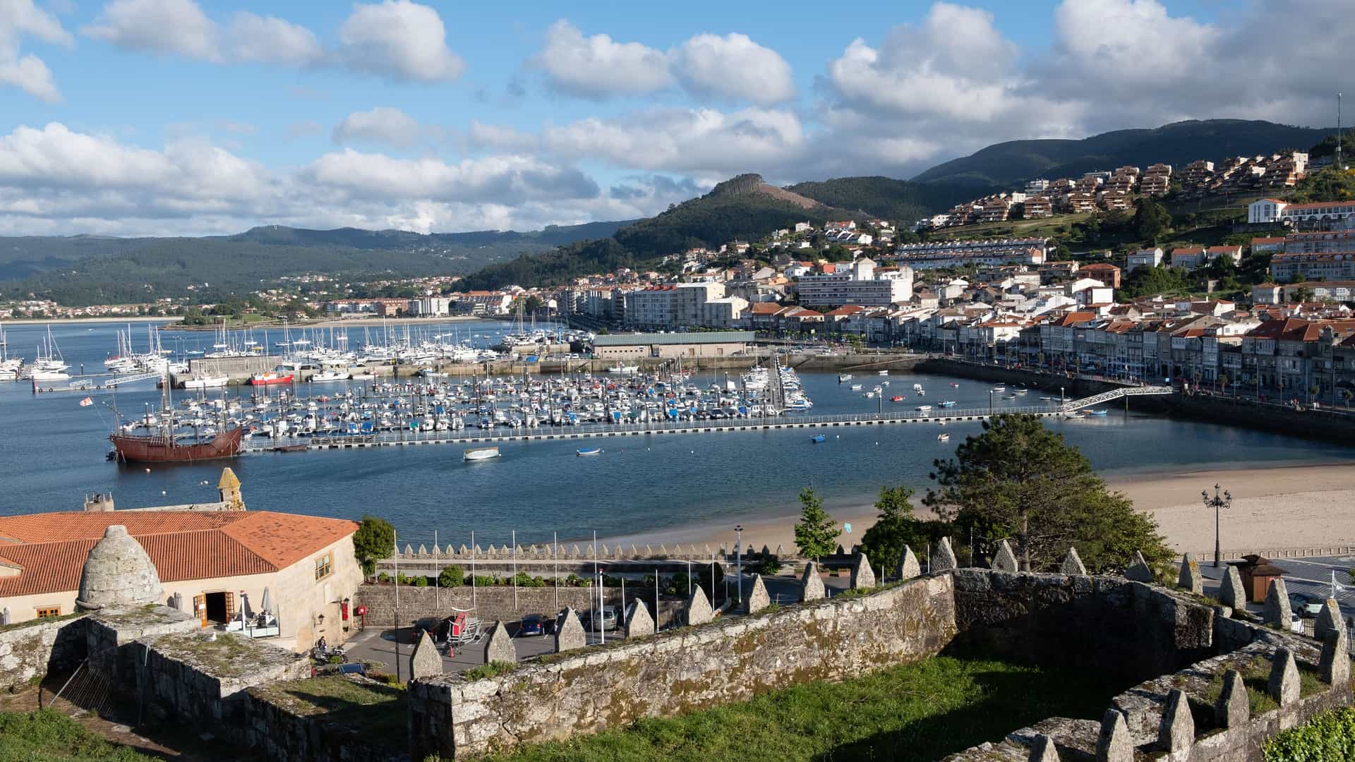 An elevated view of the picturesque harbor of Baiona, Spain, from behind a historic stone wall. The bay is filled with dozens of sailboats and yachts, while the town's white and red-roofed buildings climb the surrounding green hills.