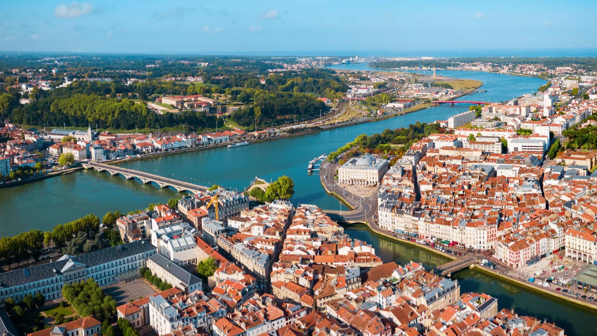 An aerial view of Bayonne, France, showing the colorful houses, river, and bridges under a clear blue sky, extending toward the ocean horizon.