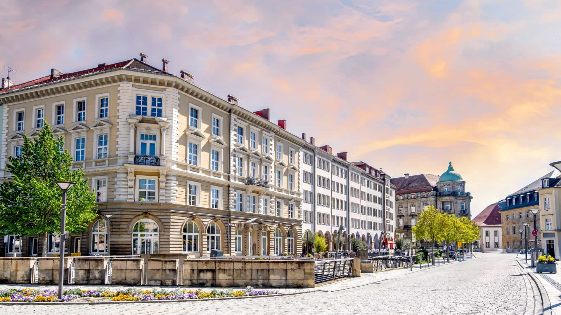 A cobblestone street view in Bayreuth, Germany, featuring grand historical buildings under a colorful, cloud-streaked sunset sky.