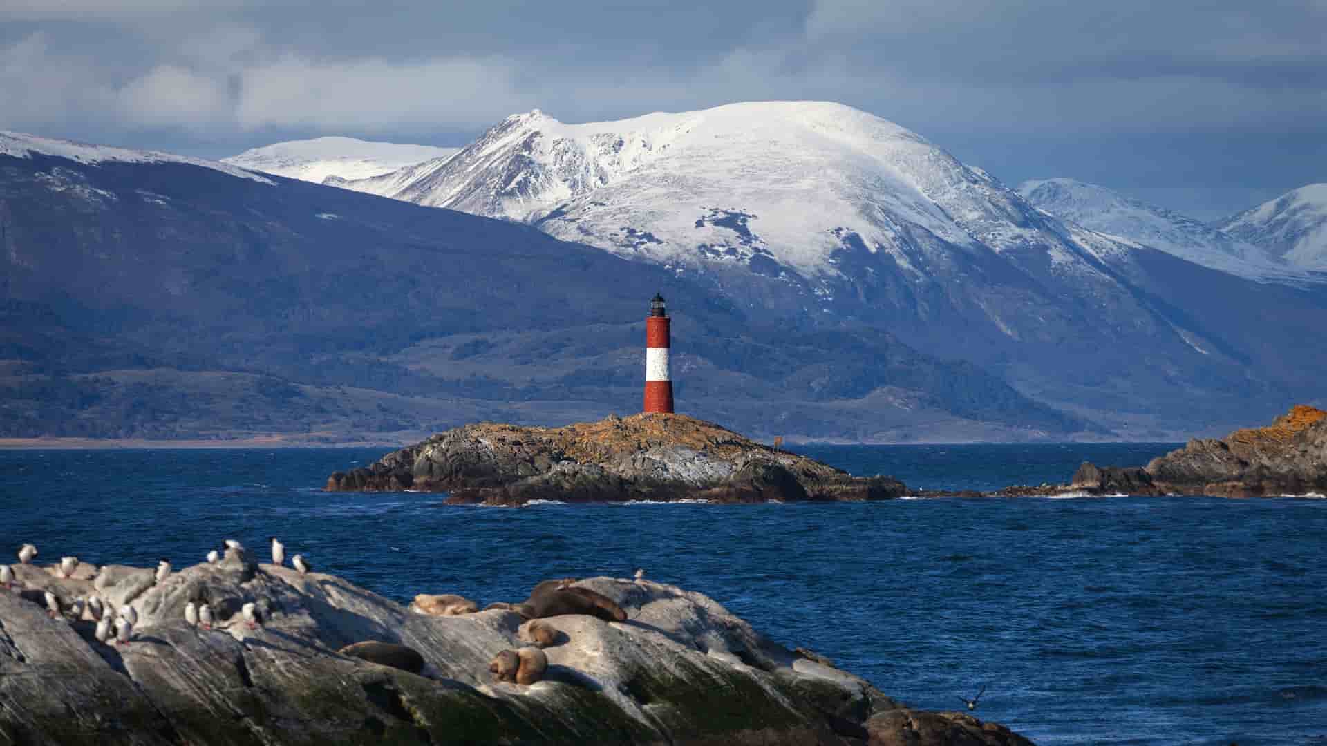 A striking image of Les Éclaireurs Lighthouse on a small rocky islet in the Beagle Channel, with seals and sea lions resting nearby and snow-capped mountains in the background.