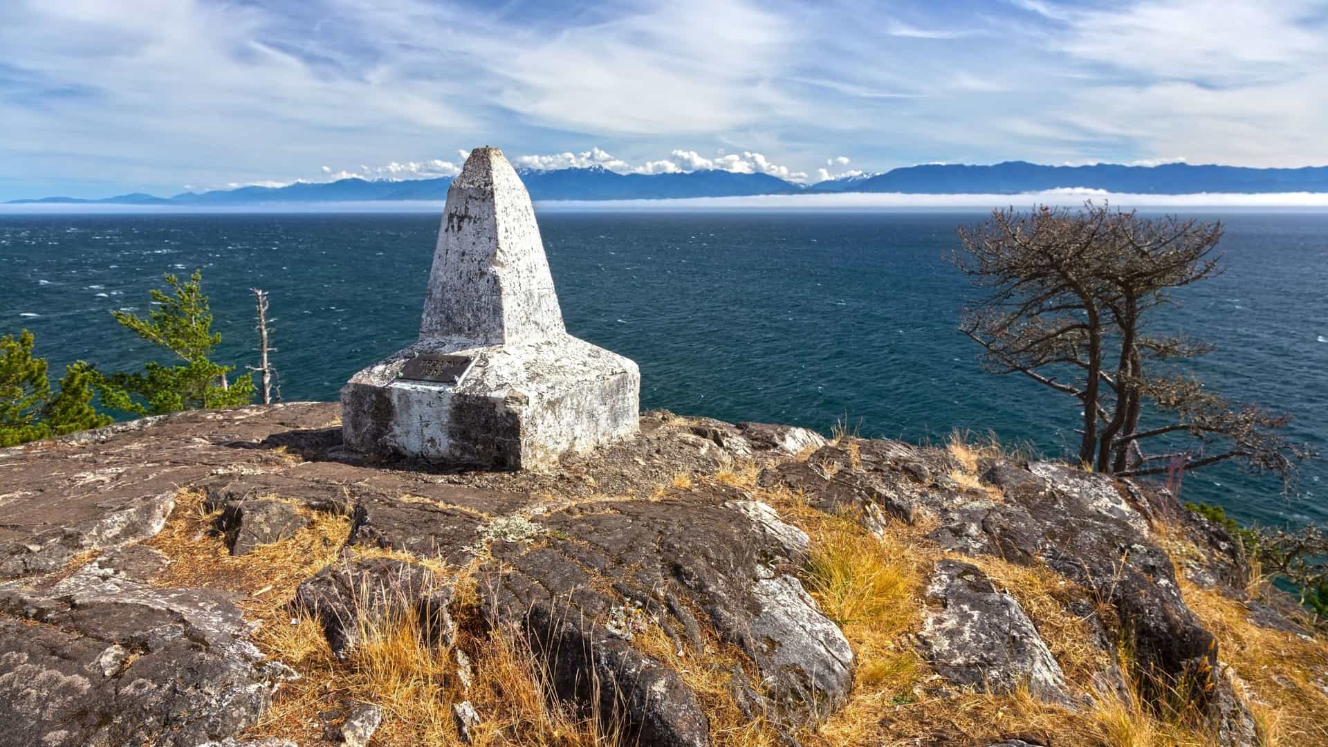 A weathered stone monument sits on a rocky cliff overlooking the vast ocean, with a view of snow-capped mountains on the distant horizon under a cloudy sky.