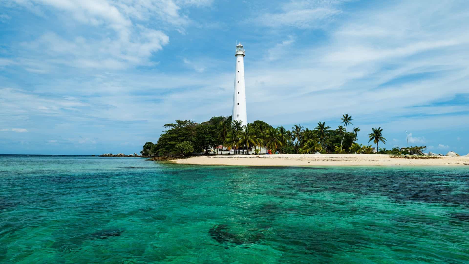 A towering white lighthouse stands on a small, lush tropical island in the Belitung Islands, surrounded by clear turquoise waters and a bright blue sky.