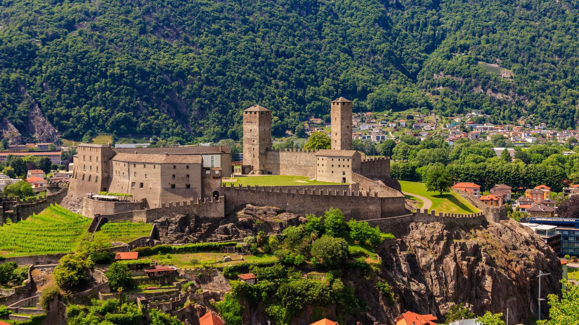 A scenic view of the Castel Grande, a large medieval castle with two towers, overlooking the city of Bellinzona in Switzerland with lush, green mountains in the background.