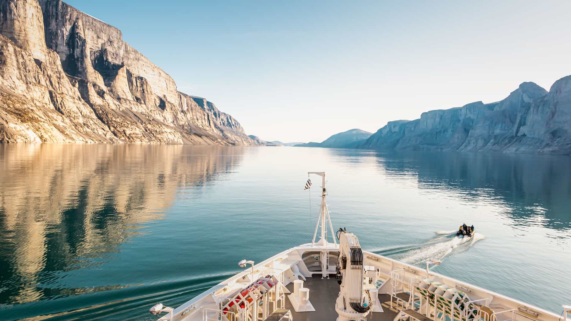 A scenic view from the bow of a ship sailing through the Bellot Strait, a narrow channel surrounded by steep, rocky cliffs with a small zodiac boat in the distance.