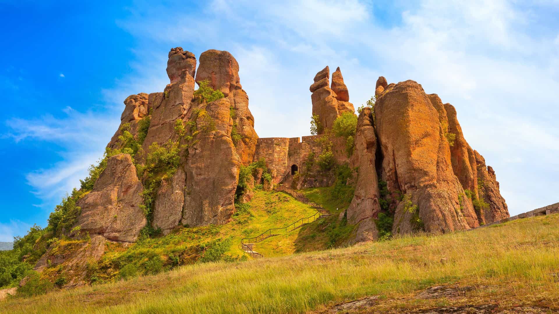 A stunning landscape shot of the Belogradchik Fortress in Bulgaria, with dramatic rock formations and the medieval fortress walls nestled between them under a bright blue sky.