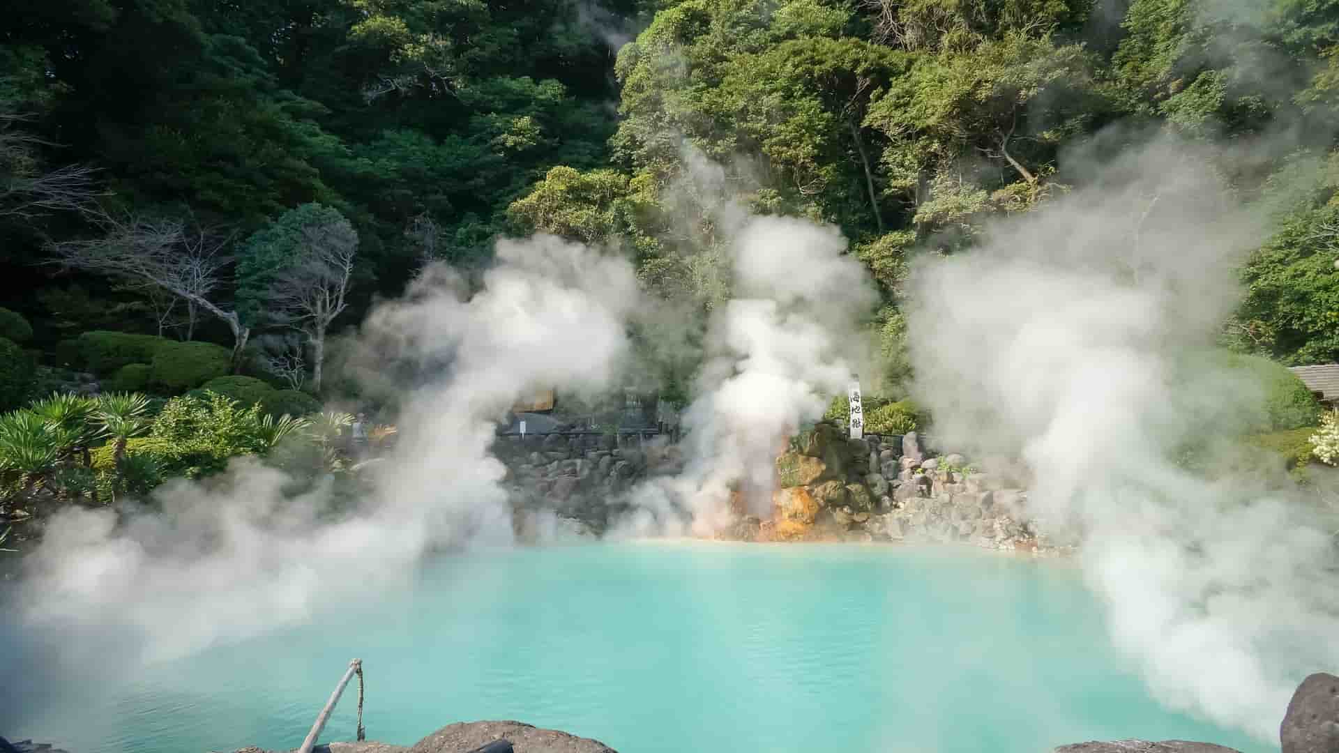 A stunning view of the Umi Jigoku ("Sea Hell") in Beppu, Japan, featuring a vibrant turquoise-blue hot spring lake emitting thick plumes of white steam, surrounded by lush green foliage.
