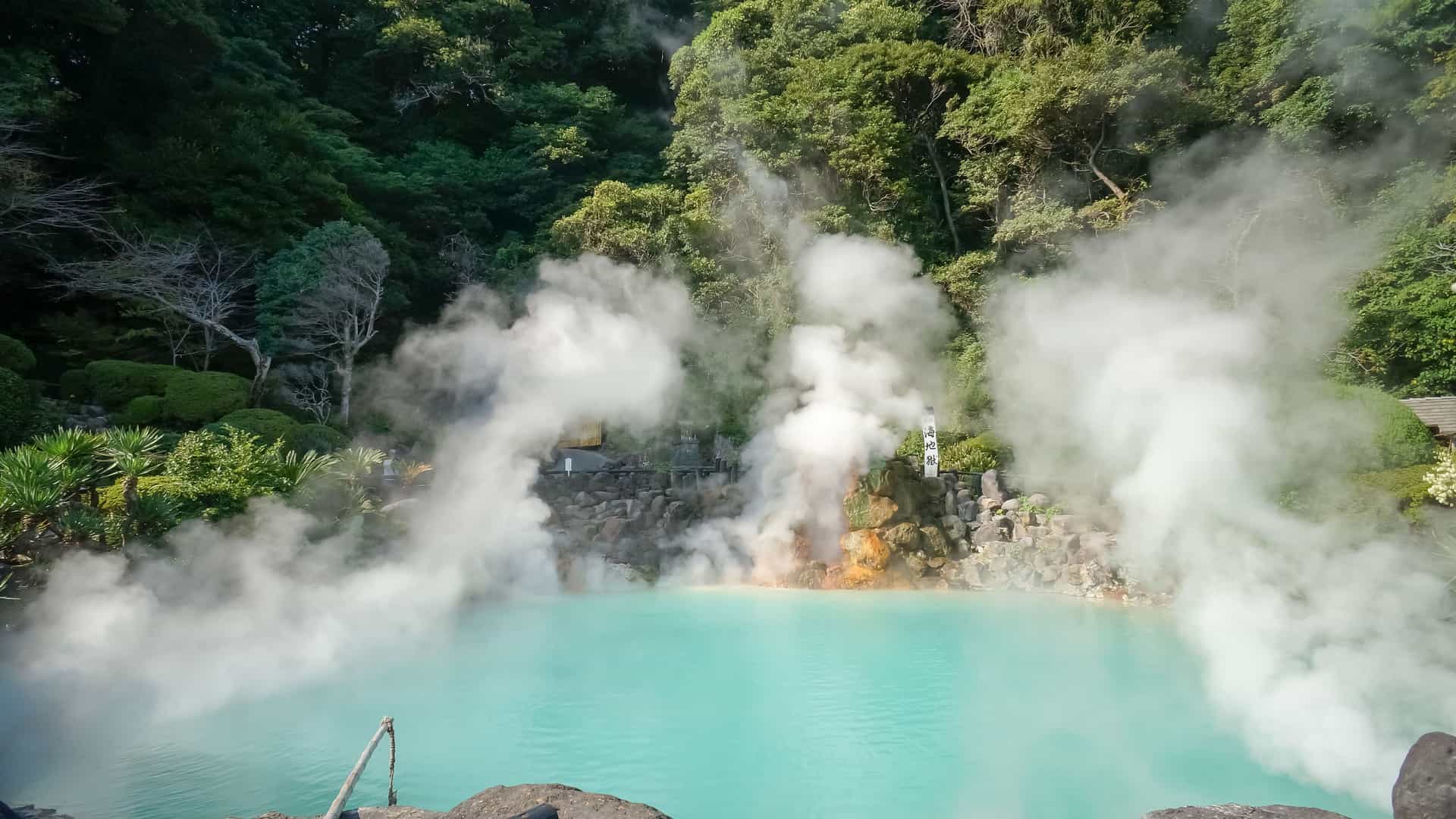 A stunning view of the Umi Jigoku ("Sea Hell") in Beppu, Japan, featuring a vibrant turquoise-blue hot spring lake emitting thick plumes of white steam, surrounded by lush green foliage.