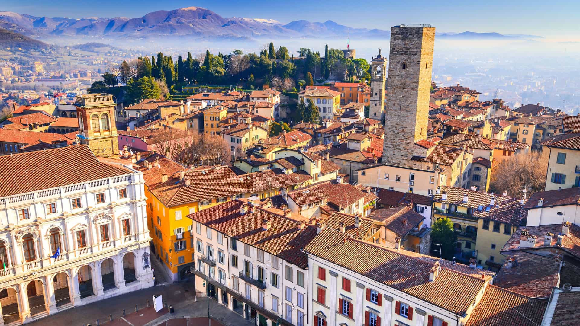 An elevated panoramic view of the historic Città Alta of Bergamo, Italy. The foreground is a dense collection of buildings with traditional red tile roofs, centered around a tall, medieval stone tower. The city spreads into the distance, with a range of snow-dusted mountains on the horizon under a clear blue sky.