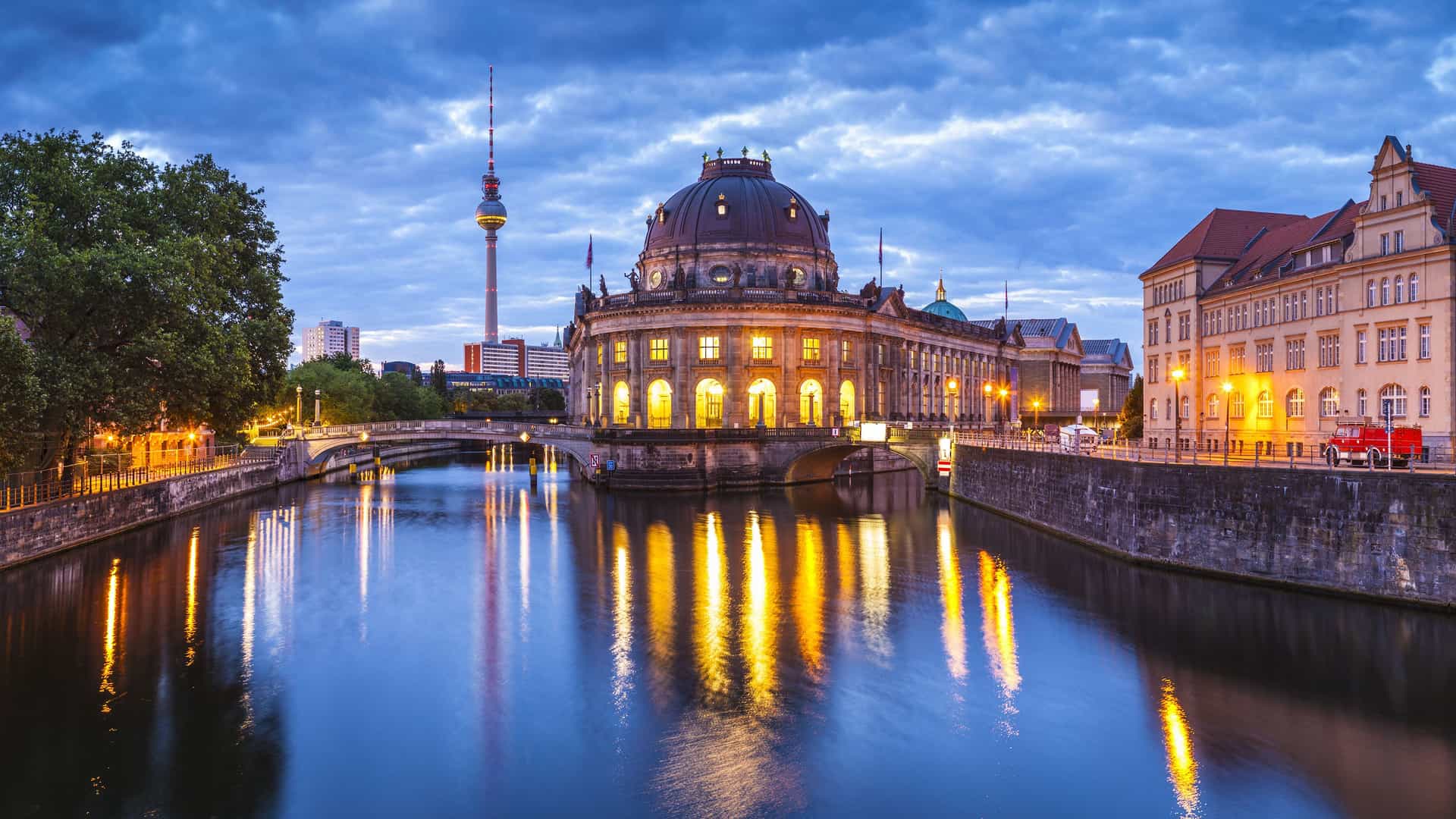 A scenic wide-angle view of the Bode Museum and the Spree River at dusk in Berlin, with the city's lights reflecting on the water.