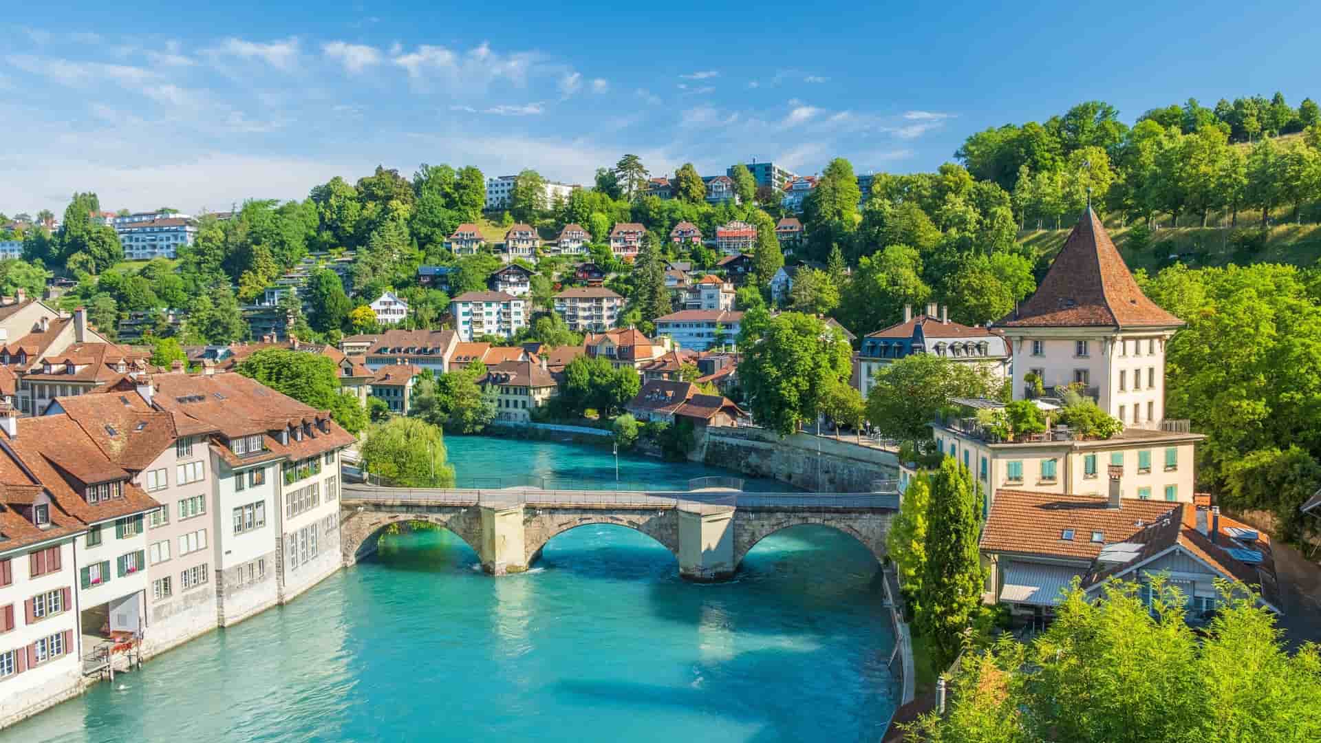 A high-angle view of the medieval city of Bern, Switzerland, with the Aare River flowing beneath the historic Untertorbrücke bridge and traditional houses lining the banks.