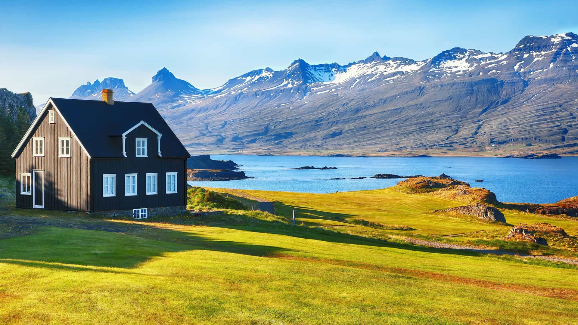 A picturesque black wooden house with white windows sits on a grassy field in the Berufjörður fjord in Iceland, with the calm blue water and snow-capped mountains in the background.