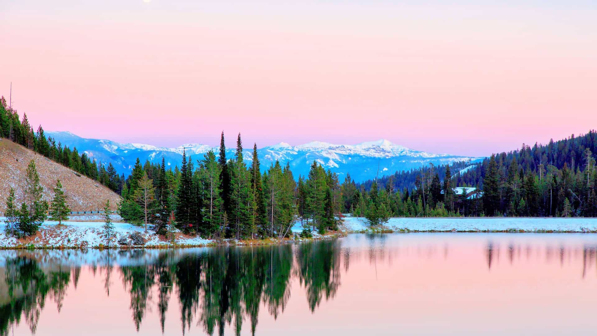 A serene landscape of a lake in Big Sky, Montana, perfectly reflecting a pastel pink and blue sky. The water's edge is lined with snow-dusted pine trees, with a range of distant, hazy snow-capped mountains visible on the horizon.
