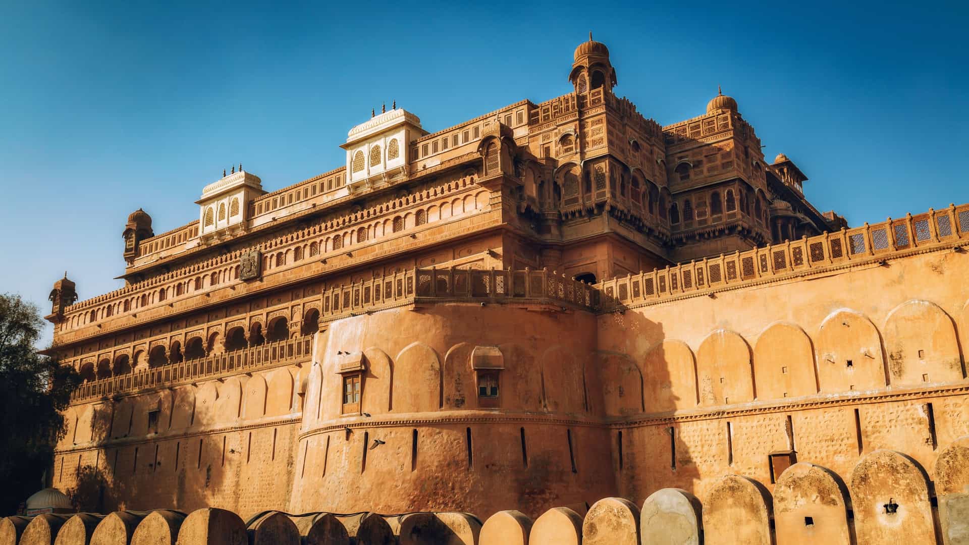 A stunning low-angle shot of the massive, historic Junagarh Fort in Bikaner, with its intricate sandstone walls and towers bathed in warm sunlight under a clear blue sky.