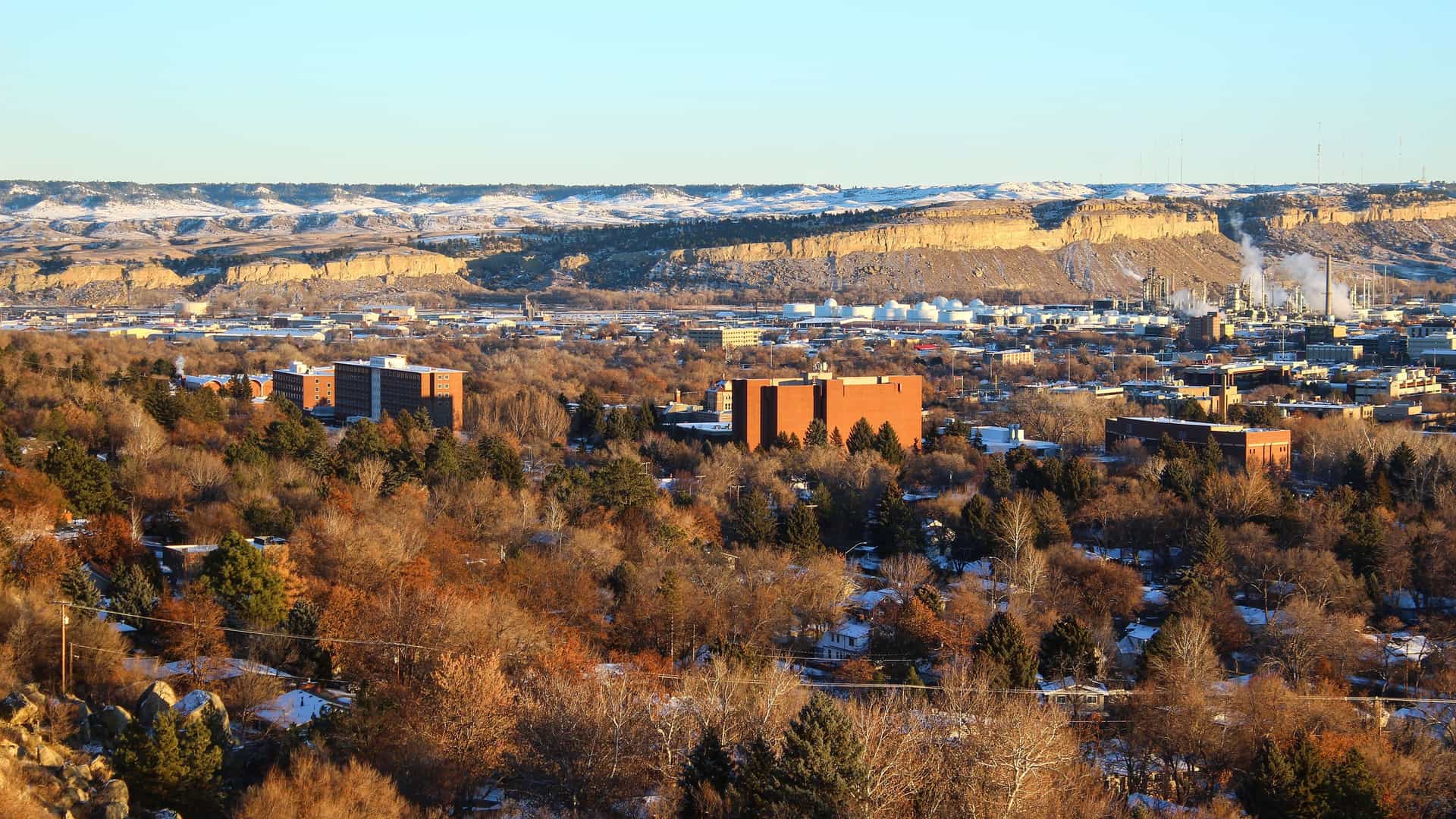 A panoramic, elevated view of Billings, Montana, during late fall or early winter, with a foreground of bare and evergreen trees and scattered buildings. The midground shows the city's downtown and industrial area, and a prominent rock formation known as the Rimrocks stretches across the background under a pale blue sky.