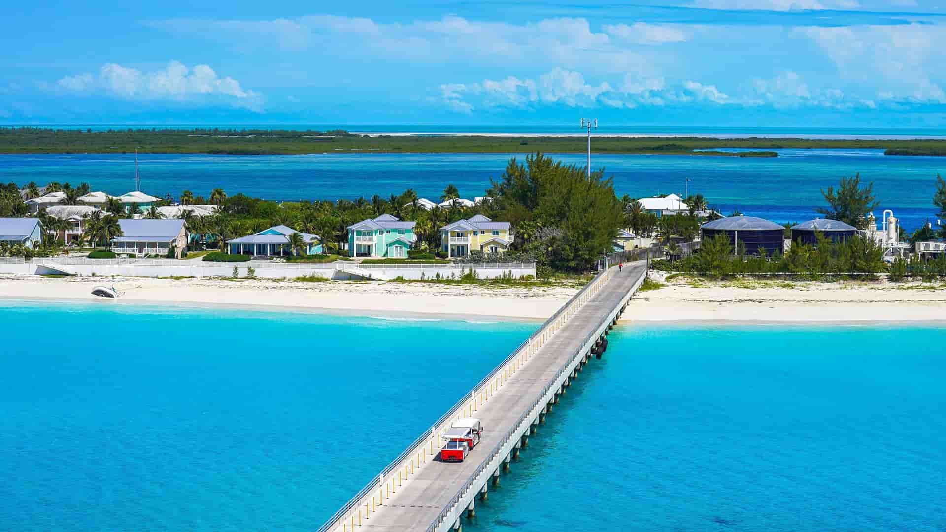 An aerial view of a long pier with a red trolley, stretching from the sandy beach of Bimini island to a collection of colorful houses, with clear turquoise water all around.