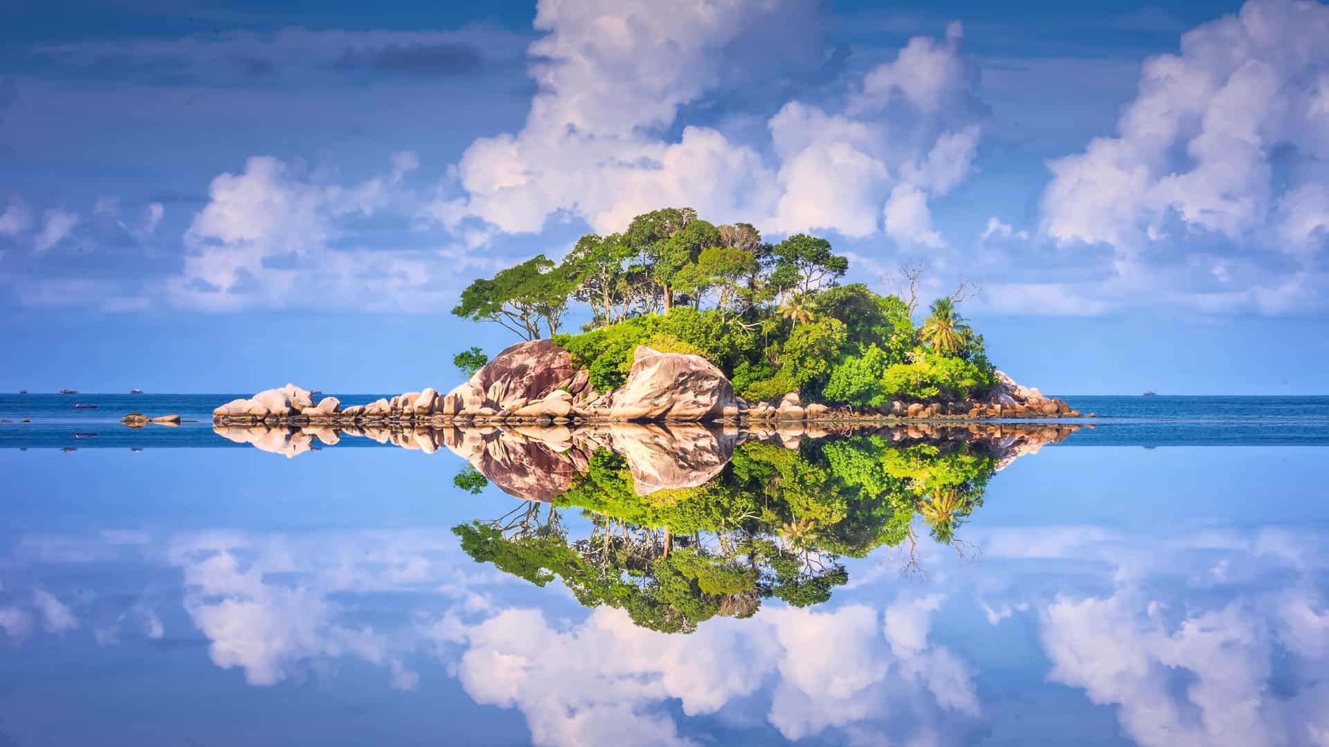 A breathtaking long exposure of a small, rocky island with lush green trees reflecting perfectly in the calm, mirror-like waters of Bintan Island, with a soft blue sky and clouds above.