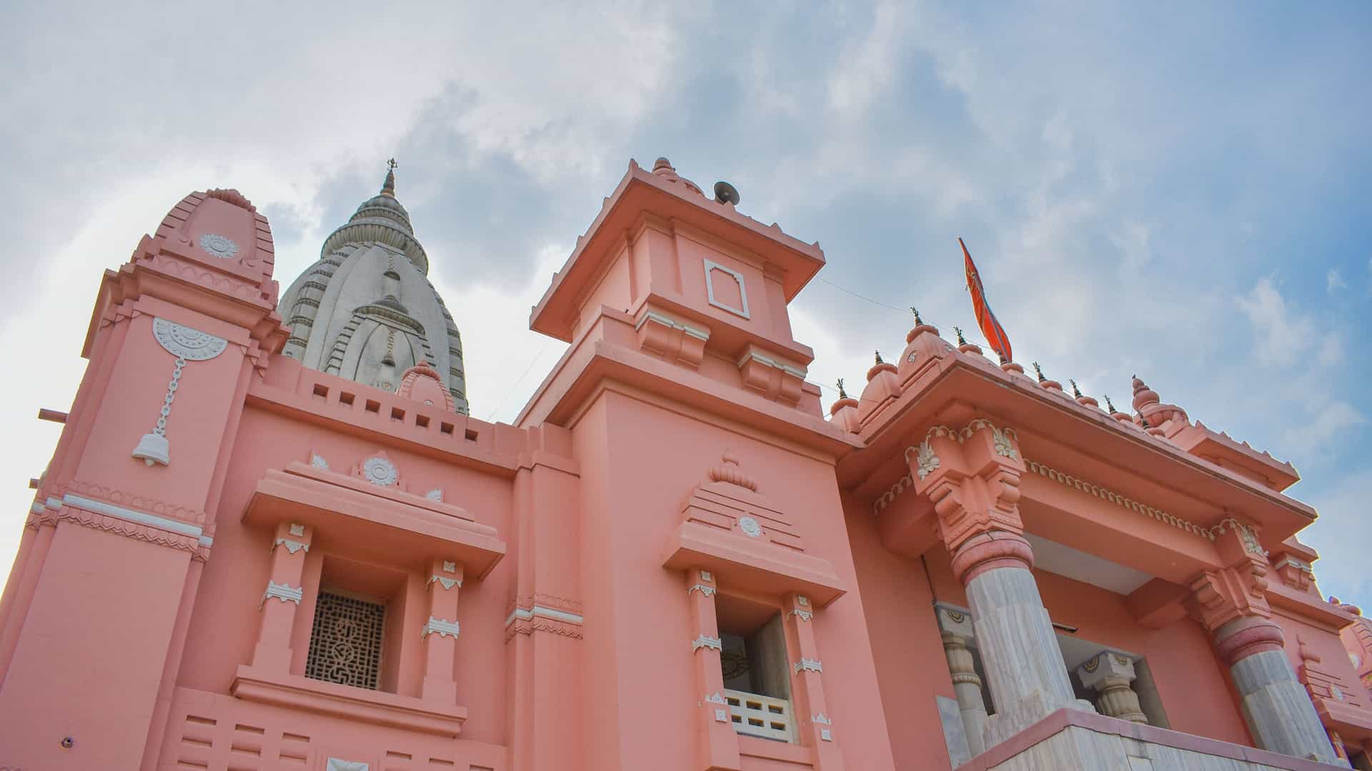 A low-angle shot of the pink-colored Biswanath Temple in Biswanath, India, with its ornate architecture, including multiple spires and columns. A prominent white dome is visible on the left side of the building, and a saffron flag flies from a spire on the right, all set against a cloudy blue sky.