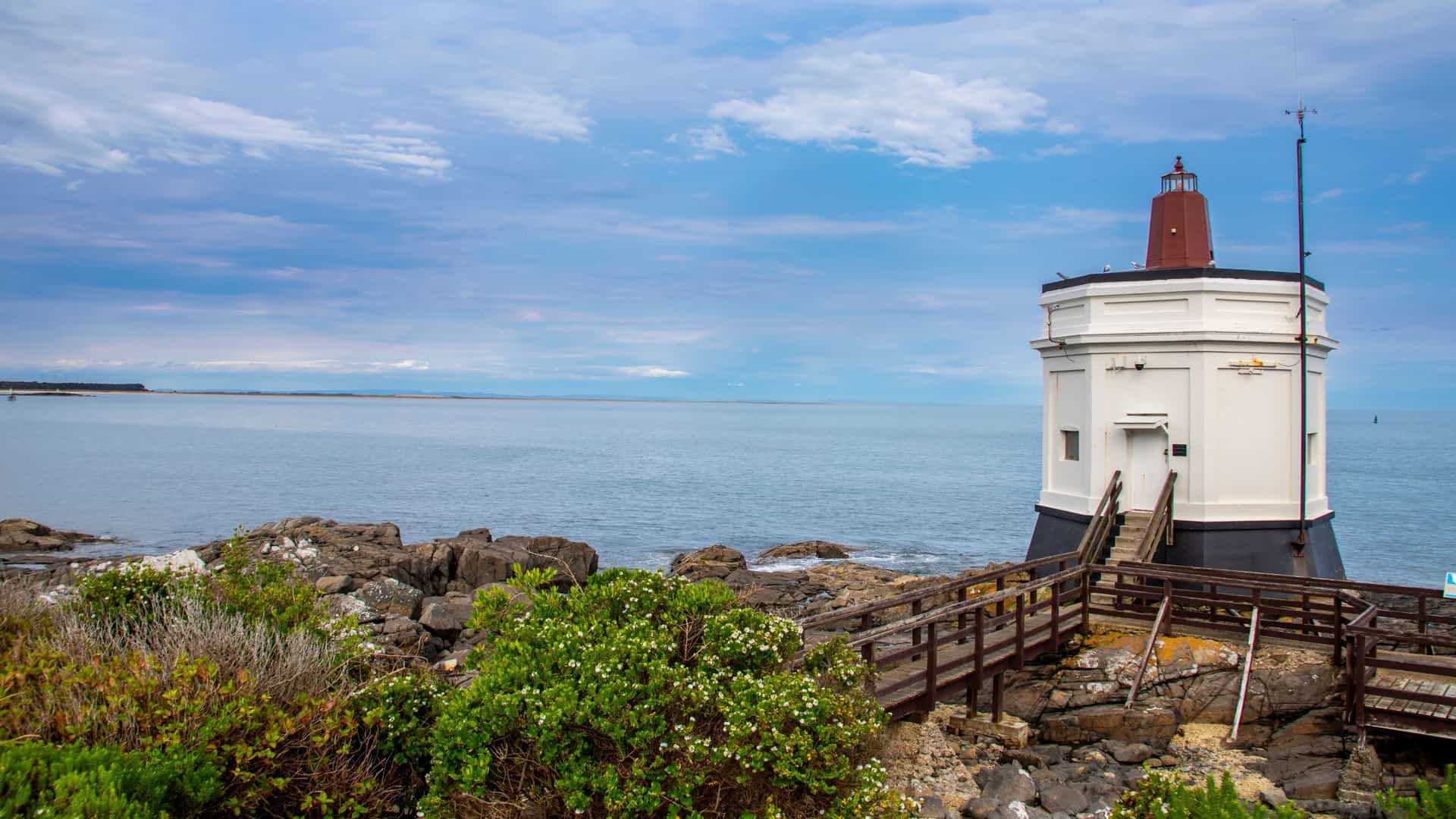 A scenic view of the iconic Bluff Lighthouse, a white and red structure with a wooden walkway, standing on a rocky shoreline overlooking the vast blue ocean under a cloudy sky.