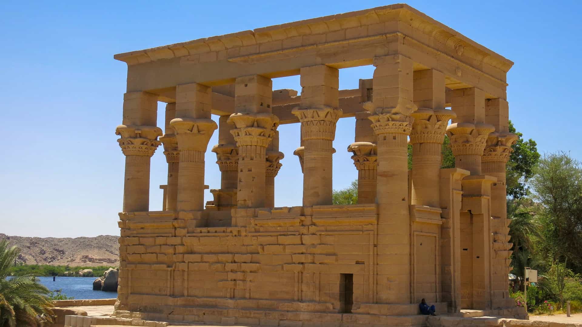 A view of the historic Trajan's Kiosk, an ancient Egyptian temple with intricately carved columns, located on the waterfront of Agilkia Island under a clear blue sky.